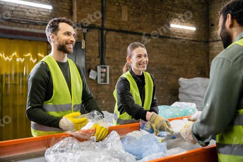 Preview: Cheerful sorters in vests and protective gloves sorting garbage and looking at blurred indian