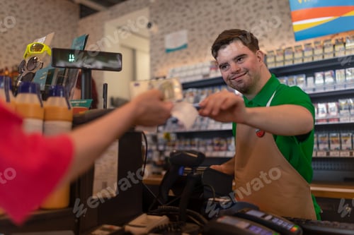 Preview: Cheerful young Down Syndrome employee taking cash payment from costumer in gas station cafe.