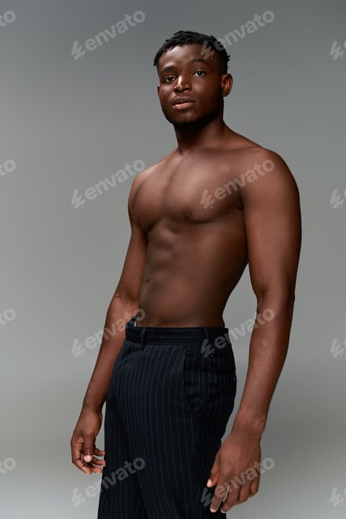 Preview: shirtless african american fitness model in black pants standing and looking at camera on grey