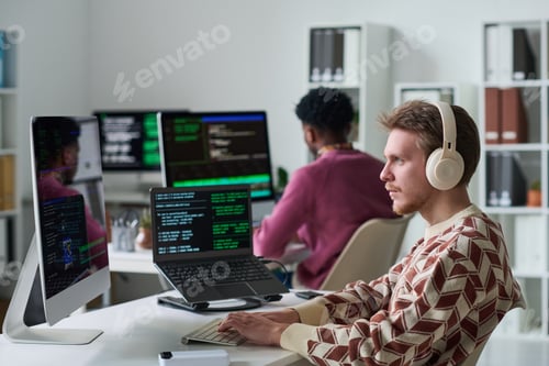 Preview: Young Man Wearing Headphones Coding Beside Black Man in Office