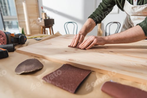 Preview: Cropped view of repairman sanding wooden board near sandpaper in workshop