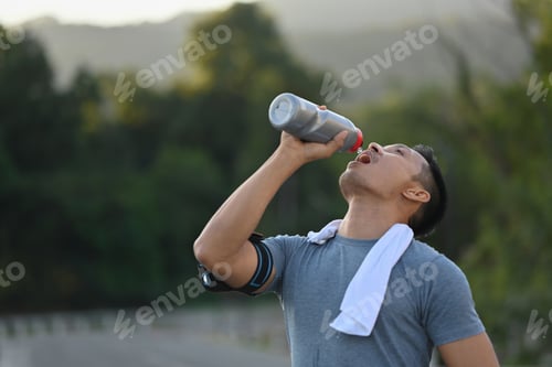 Preview: Young athletic man drinking water from a bottle, taking a break from workout in the park.