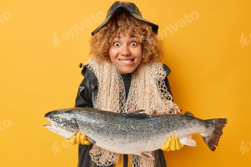 Preview: Positive fisherwoman with curly hair bites lips holds huge caught salmon carries fishnet around neck