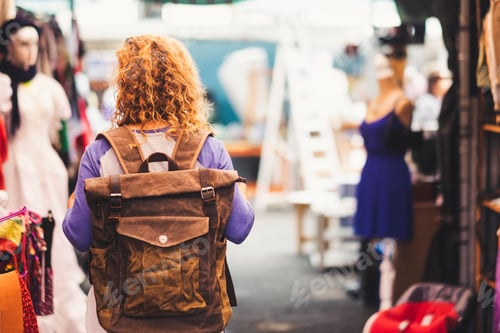 Preview: Blonde curly hair woman backpacker traveler viewed from rear at used market enjoying