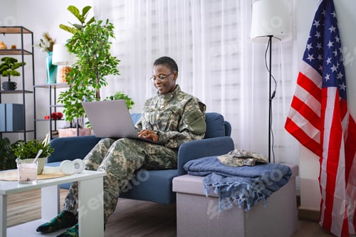 Preview: Female soldier sitting on sofa using laptop with United States flag in background