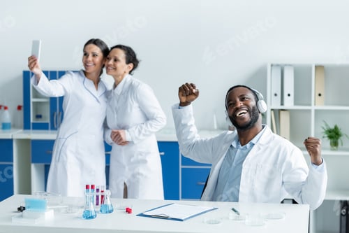 Preview: African american scientist in headphones showing yes gesture near blurred colleagues with smartphone