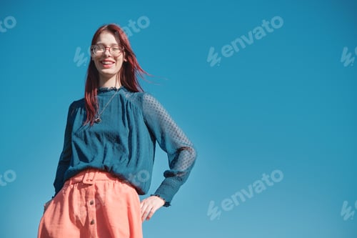 Preview: Smiling teenage girl with red hair and glasses wearing blue blouse and coral skirt.