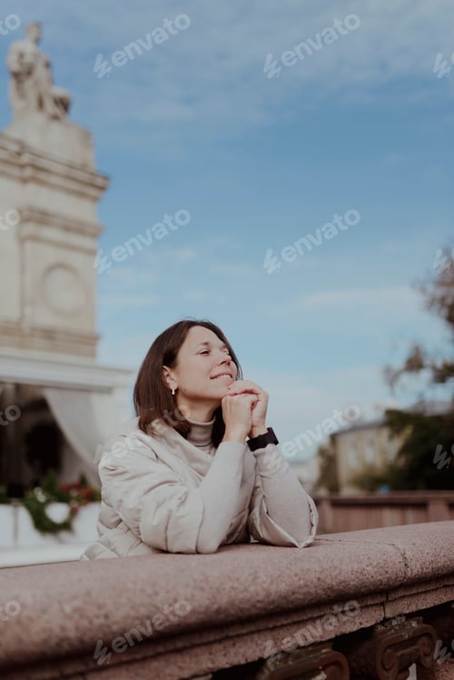 Preview: A beautiful, thoughtful woman in beige clothes is leaning on the stone railing.