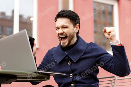 Preview: Successful young man celebrating on laptop sitting in cafe outdoors in the city
