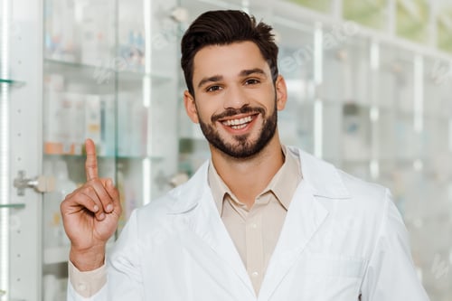 Preview: Handsome pharmacist smiling at camera and pointing with finger in pharmacy