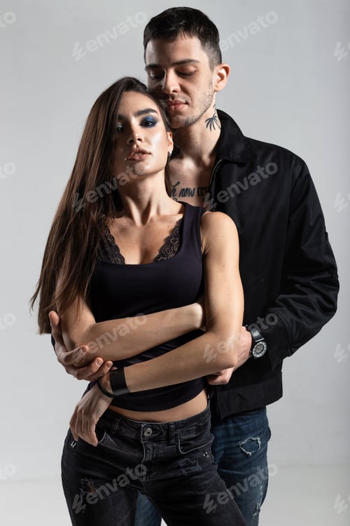 Preview: Cocky guy in black jacket on a naked torso is hugging from behind a brunette girl dressed in a black
