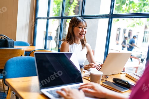 Preview: Cheerful Asian woman and Caucasian colleague working on laptops in a bright Chiang Mai cafe