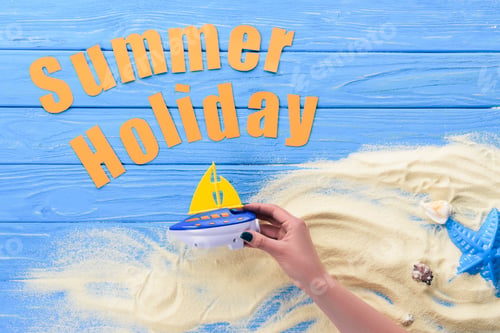 Preview: Female hands with toy ship by Summer holiday inscription on blue wooden background