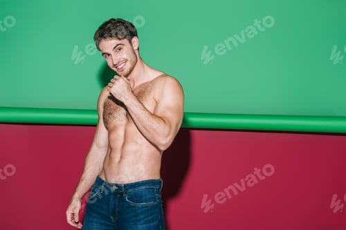 Preview: sexy shirtles man in denim jeans smiling at camera on green and red background