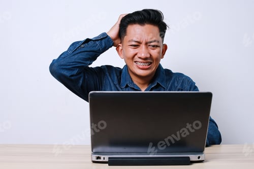 Preview: Confused Asian Adult Male Touching His Head While Using Laptop