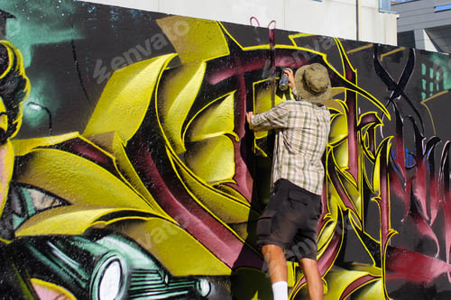 Preview: Rear view of a young graffiti artist spray painting on a concrete wall on the street