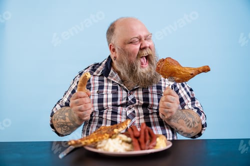 Preview: Hungry man with overweight eats chicken leg at table with fat food on blue background
