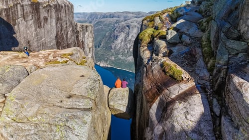 Preview: Two hikers sit on the edge of Kjeragbolten Pulpit Rock in Norway, enjoying the stunning panoramic