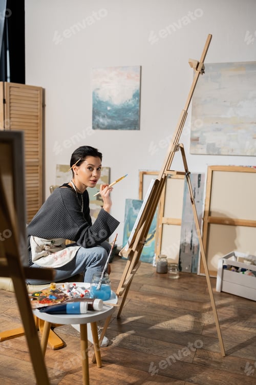 Preview: Brunette artist holding paintbrush and looking at camera near canvas on easel in studio