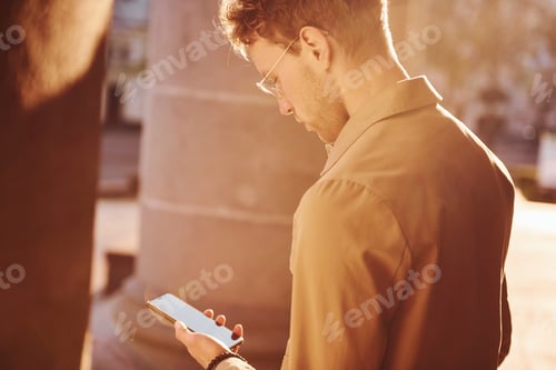Preview: Holding phone in hand. Elegant young man in formal classy clothes outdoors in the city
