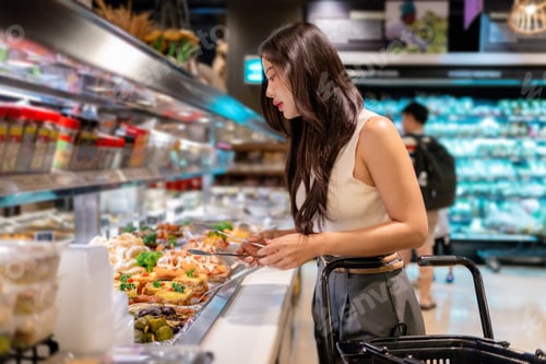 Preview: Customer using tongs to select food from display of ready meals inside grocery store