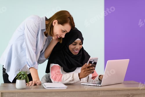 Preview: afro girl with a hijab and a European woman use a cell phone and laptop in their home office