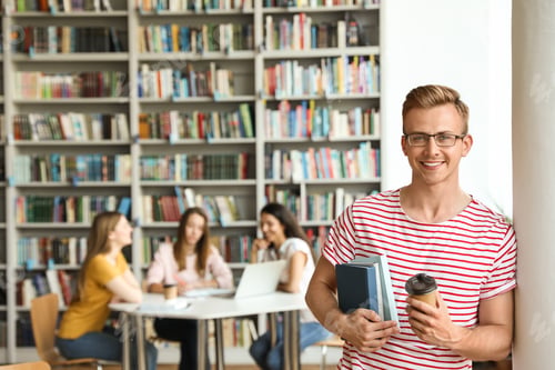 Preview: Young man with books and drink in library. Space for text