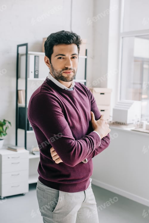 Preview: handsome designer in burgundy sweater standing with crossed arms and looking at camera in office