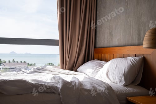Preview: Cozy bed with white sheets and pillow in sunlit room by large window overlooking the sea