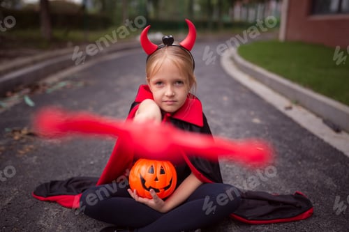 Preview: Happy Little Caucasian Girl in costume of devil with red horns for Halloween is sitting on road