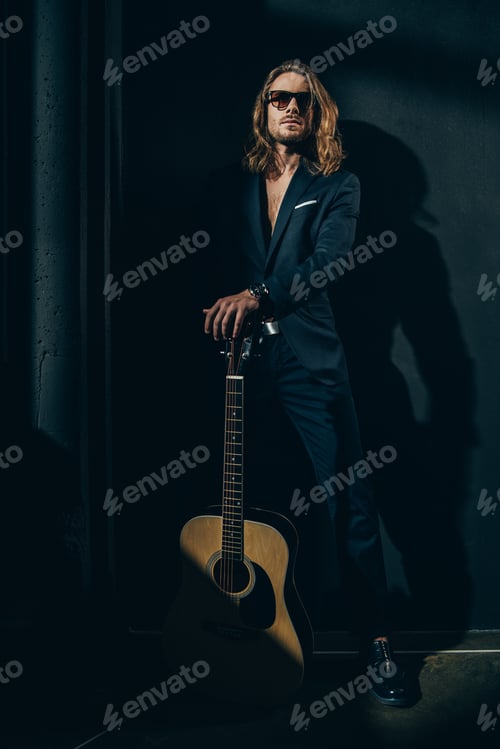 Preview: Handsome long haired man in sunglasses and stylish suit posing with acoustic guitar