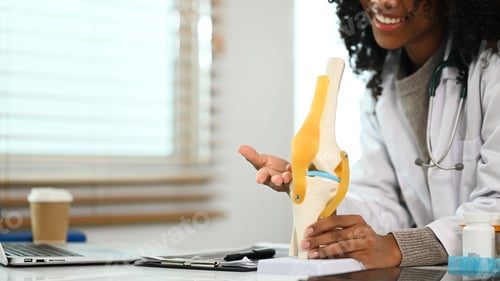 Preview: African American female doctor showing knee joint model during online consultation with patient.