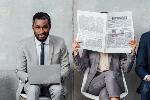 Preview: smiling african american businessman using laptop while businesswoman reading newspaper in waiting