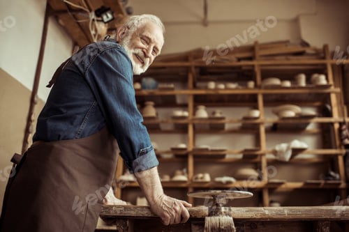 Preview: Side view of senior potter in apron standing and leaning on table against shelves with pottery goods