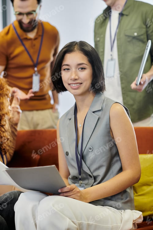 Preview: professional headshot of young smiling woman with documents near blurred colleagues, team lead