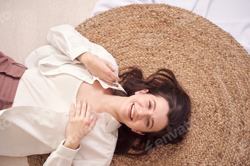 Preview: Joyful woman relaxing on woven rug smiling in stylish outfit indoors