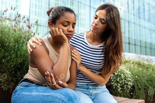 Preview: Young woman consoling and comforting upset African American female friend in park outdoors.