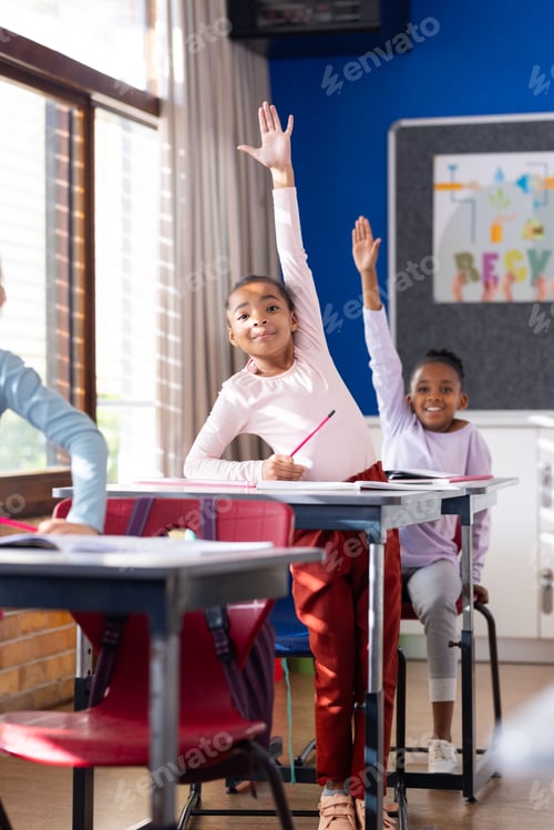 Preview: In school, diverse students raising hands while sitting at desks in classroom