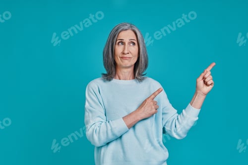 Preview: Mature beautiful woman making a face and pointing copy space while standing against blue background