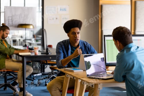 Preview: Diverse male colleagues in discussion at desks using laptops in office