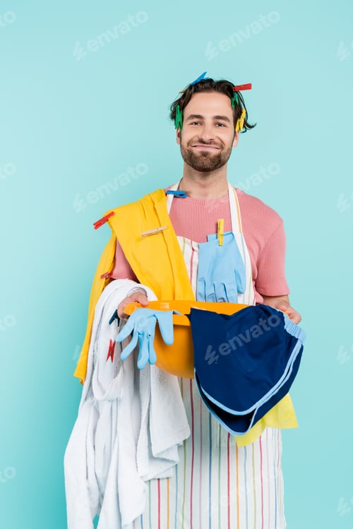 Preview: Smiling man in clothespins holding wash bowl with clothes isolated on blue
