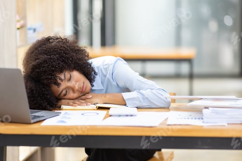 Preview: Exhausted millennial African woman sleeping on her office desk, next to laptop and documents, tired