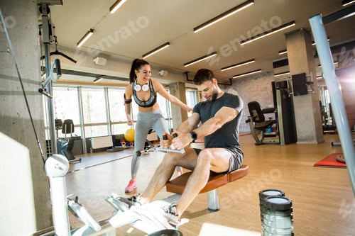 Preview: Young man using seated row machine in the gym with support of female coach