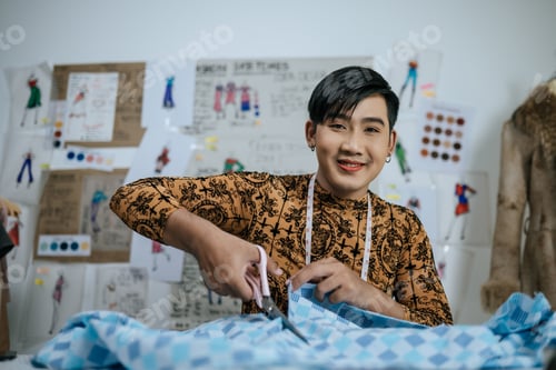 Preview: Portrait of Asian young male tailor cutting piece of cloth at table in fashion studio.
