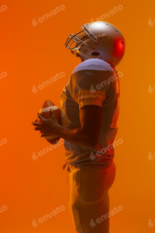 Preview: African american male american football player holding ball with neon orange lighting