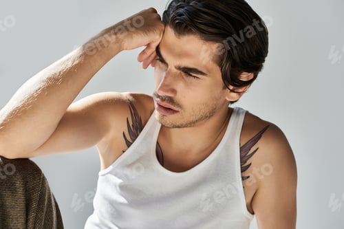 Preview: Stylish young man with tattoos in a tank top poses thoughtfully against a grey background