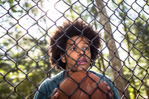 Preview: Portrait of young man holding basketball through wire fence