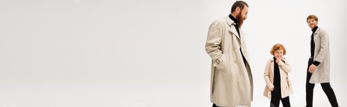 Preview: smiling redhead kid in trench coat posing with hand in pocket near father and granddad on light grey