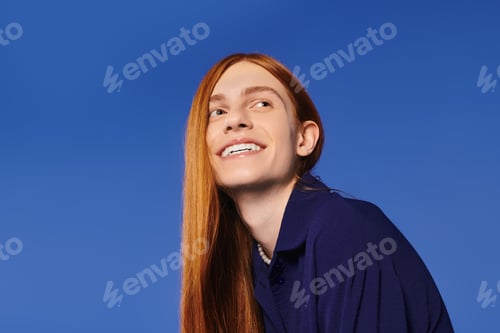 Preview: Stylish young man with long red hair smiles brightly against a vibrant blue background