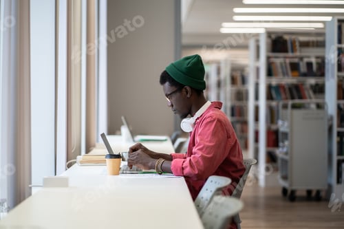 Preview: Focused African American student guy sitting at desk with laptop using online library database